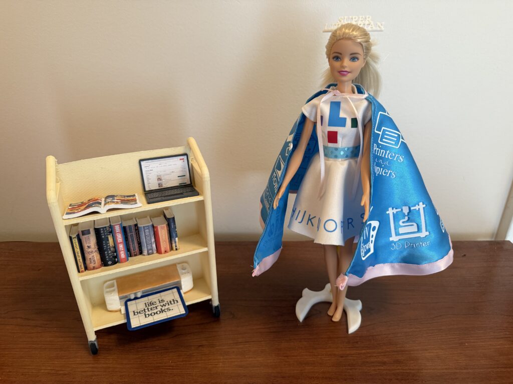 A Barbie with a blue cape and ORL logos standing beside a bookshelf.